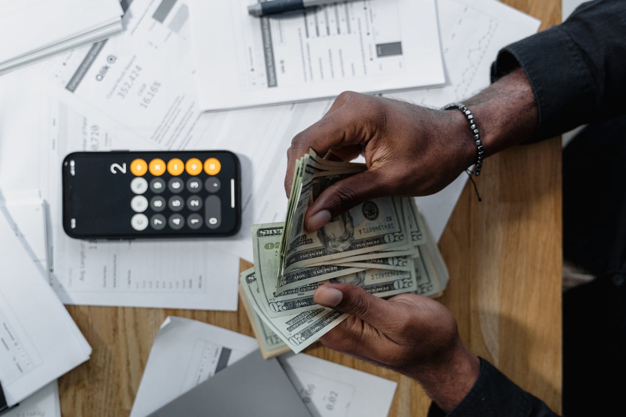 Person counting dollar bills over documents with a smartphone calculator on the desk.
